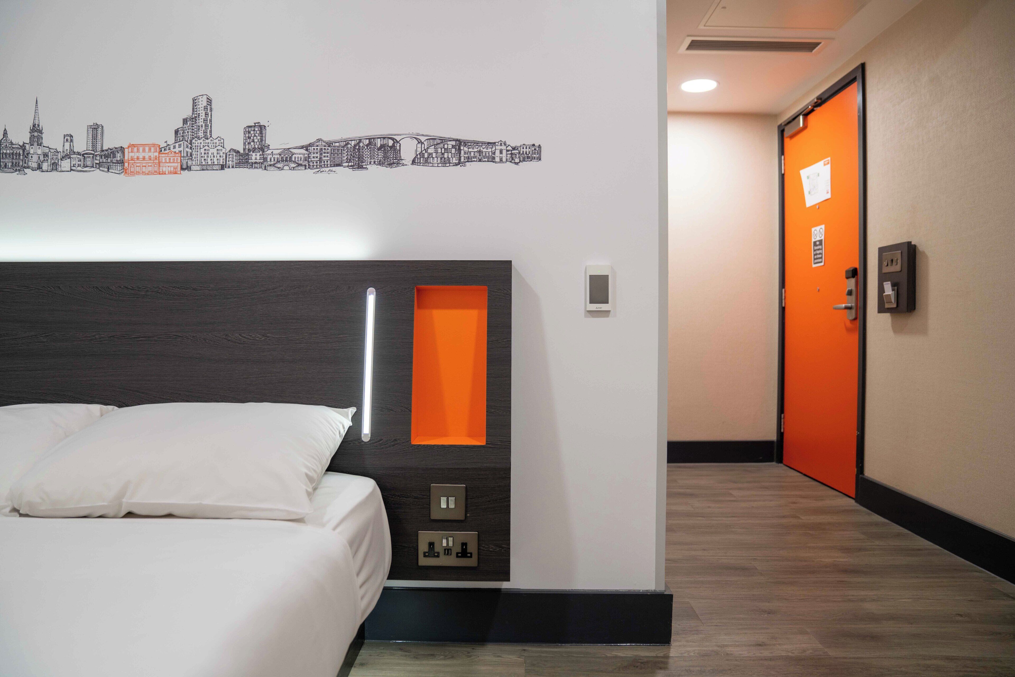 A hotel room with a bed, a cityscape wall art, an orange accent wall, and a door with an emergency exit sign.