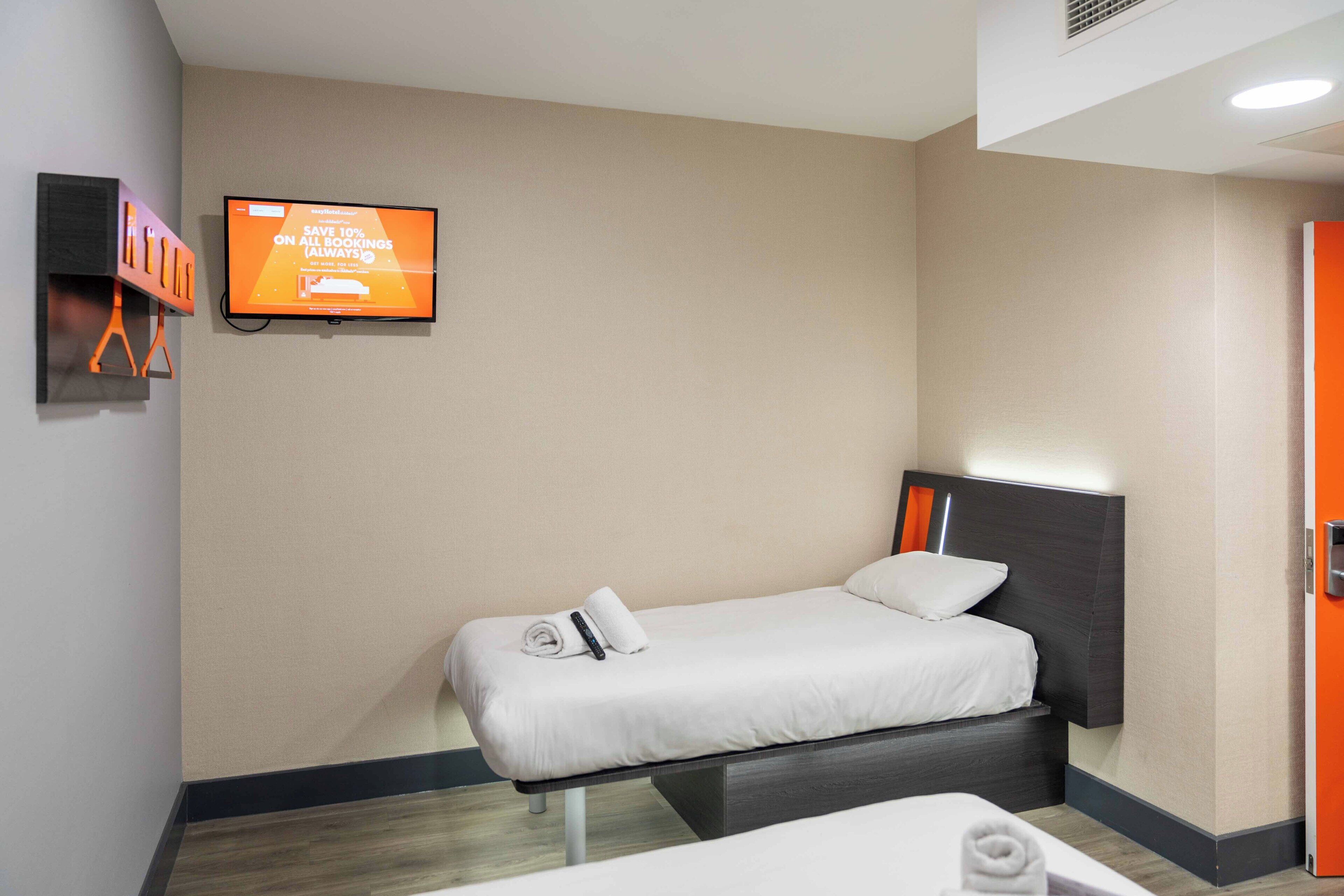 Een enkel bed met hoofdeinde, een aan de muur bevestigde televisie en een deur aan de rechterkant.