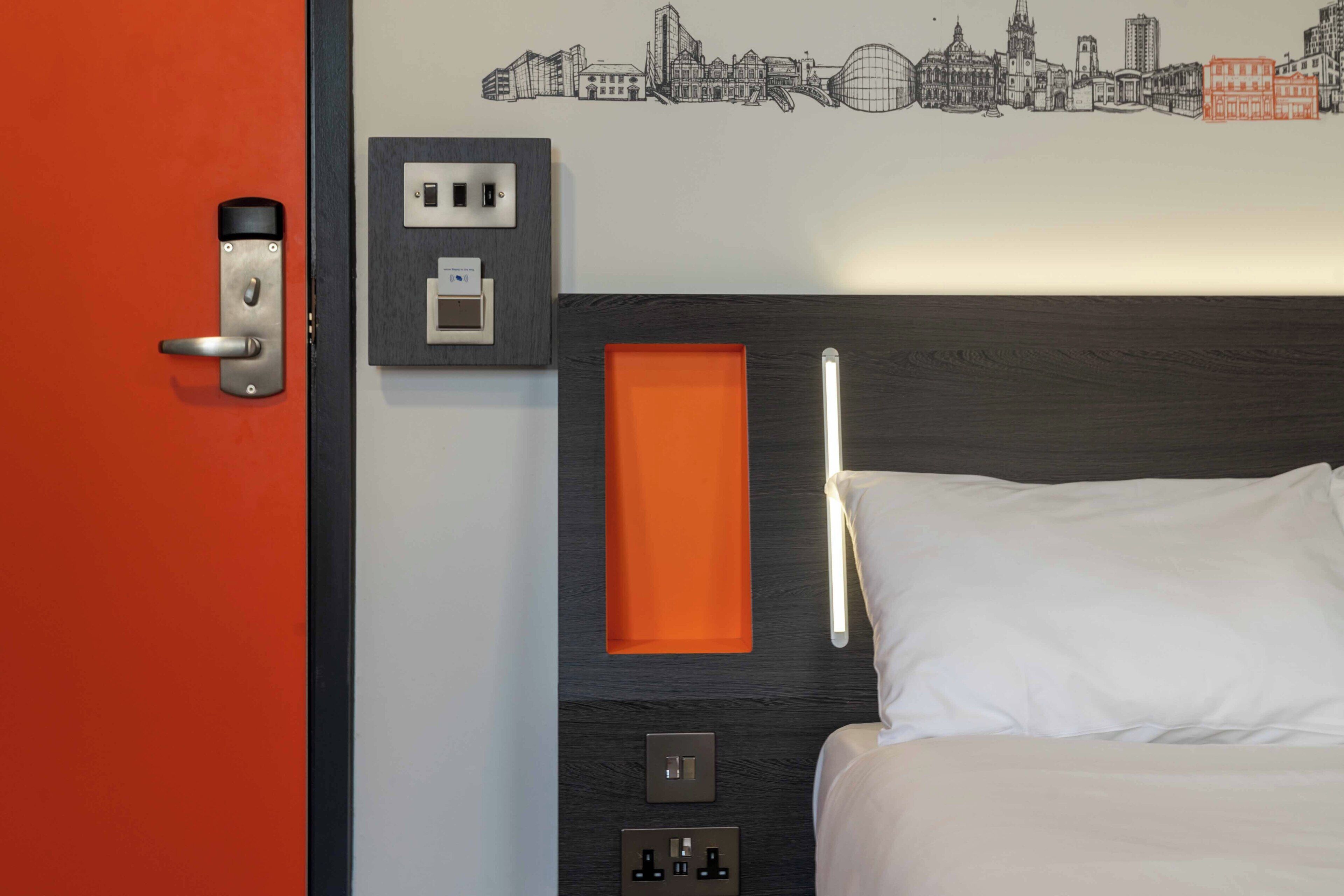 A hotel room with a bed, a wall socket, a power outlet, and a door with a handle.