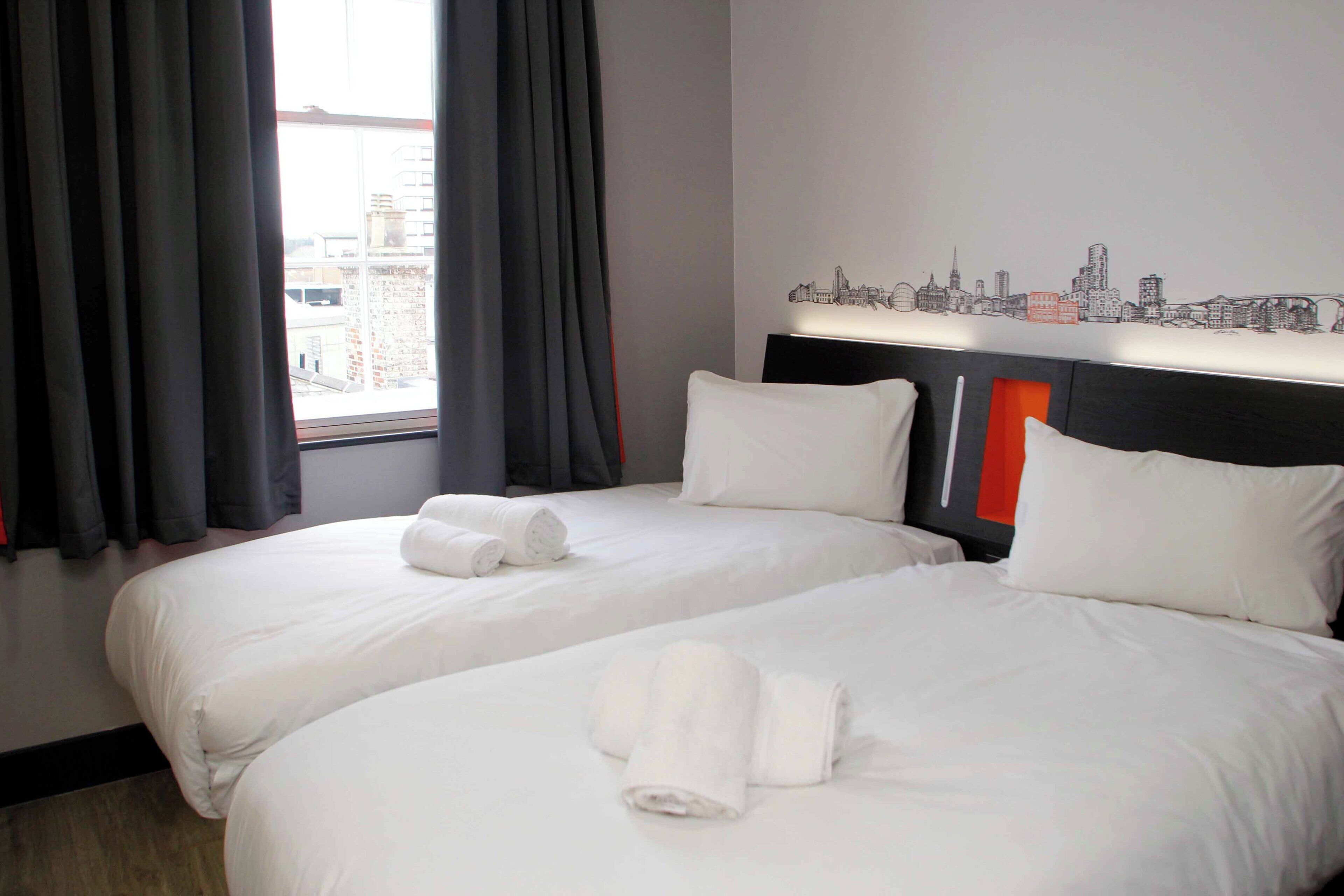 A hotel room with two beds, a city skyline mural, and a window with curtains.