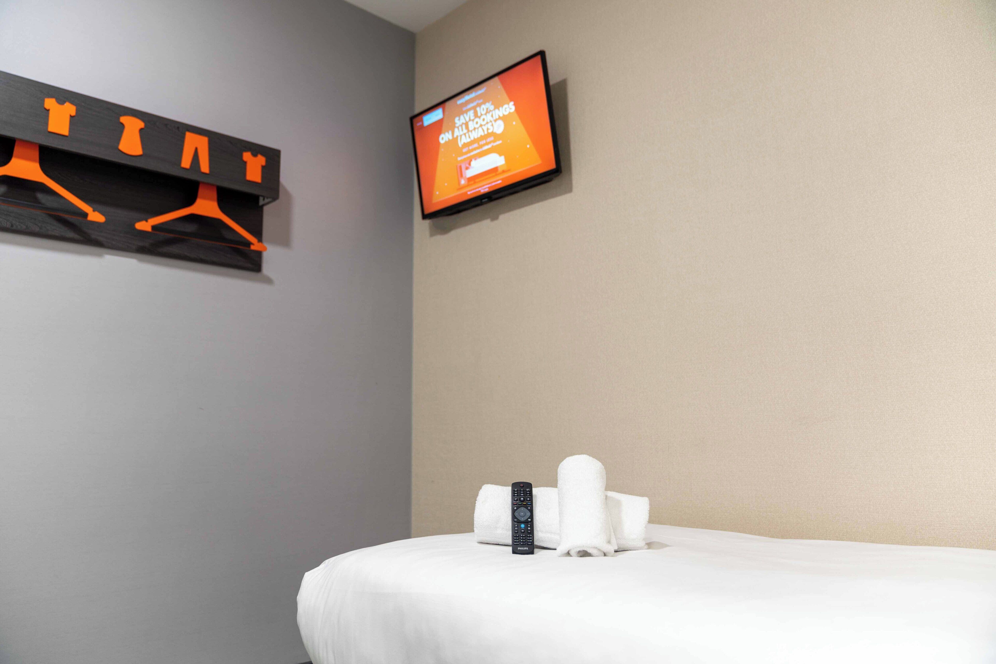 A hotel room with a bed, a TV mounted on the wall, and a towel rack with hangers.