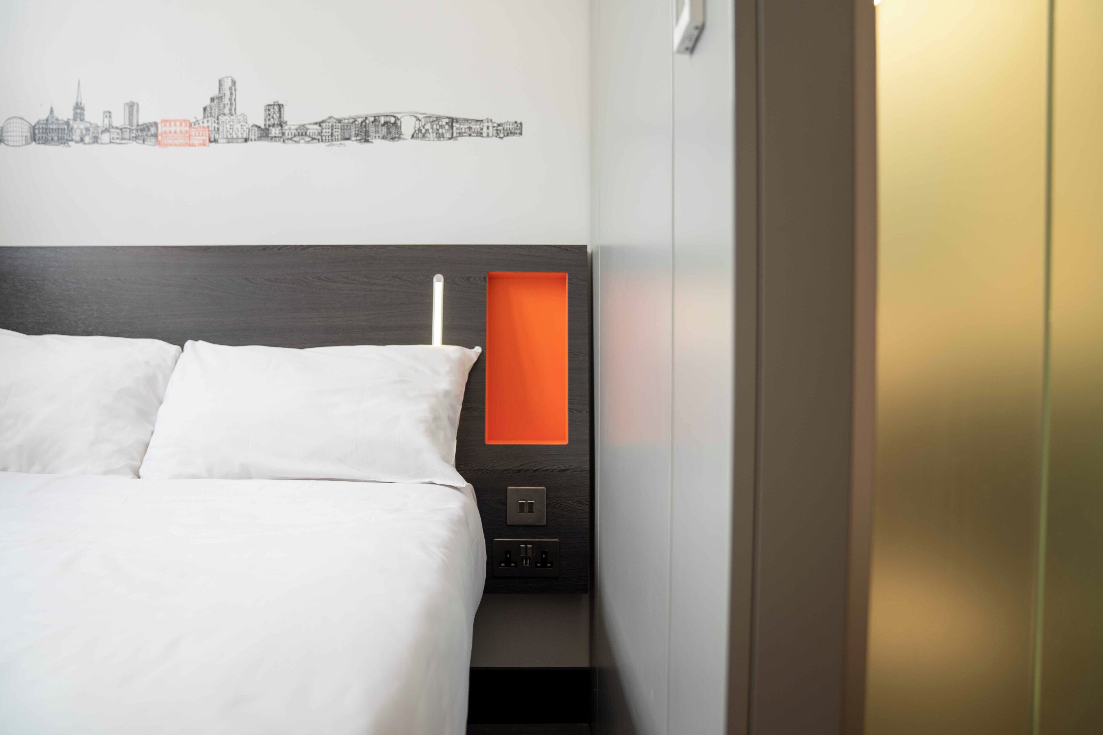 A hotel room with a bed, a cityscape wall art, a nightstand with a lamp, and a door.