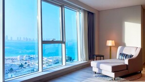 Habitación de hotel moderna con amplios ventanales desde el suelo hasta el techo que ofrecen vista a la ciudad, un sofá cómodo, una mesita auxiliar pequeña y una lámpara.