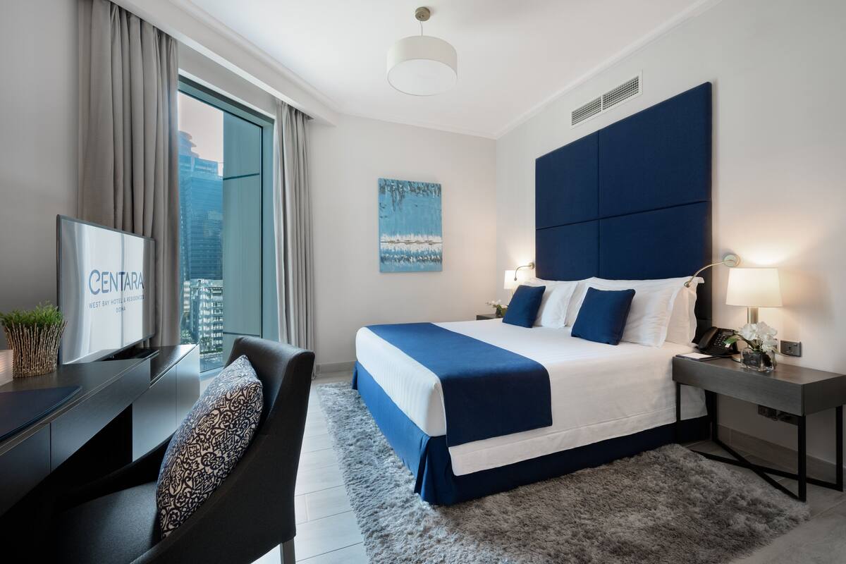 Una habitación de hotel moderna con una cama grande, un escritorio y un televisor mostrando el nombre del hotel.
