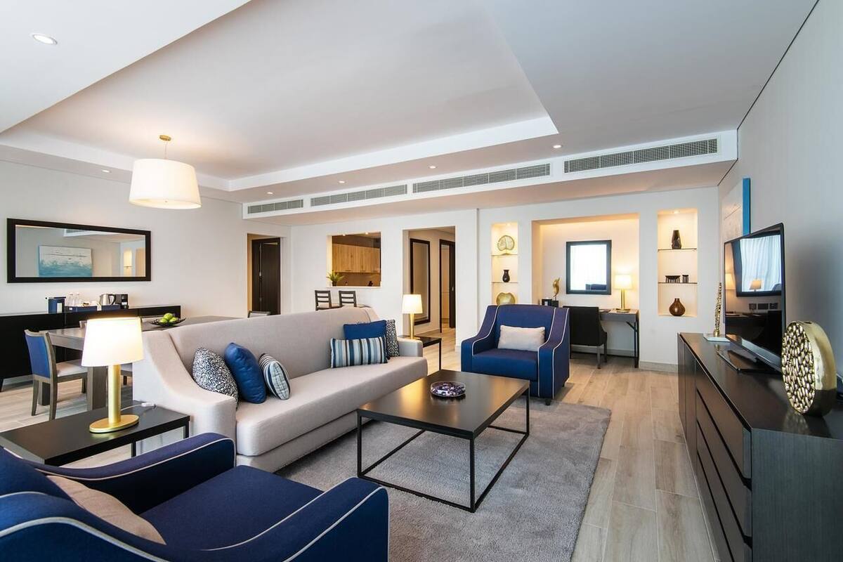 Una sala de estar moderna con un sofá, sillones, una mesa de centro y un televisor. Se ve un comedor con una mesa y sillas al fondo.