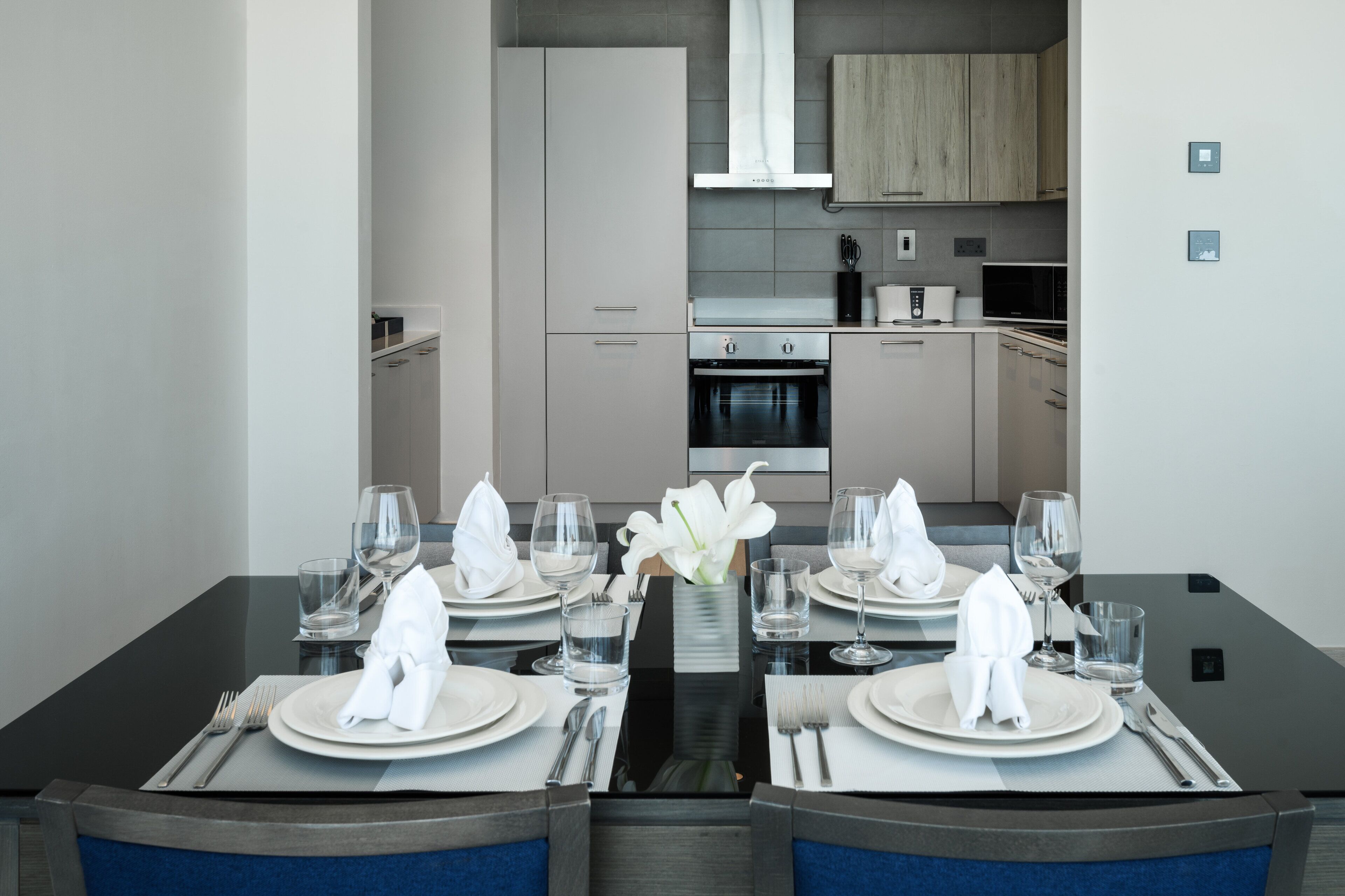 A modern dining area with a black table set for four, including plates, glasses, and cutlery. A vase with white flowers is centered on the table.