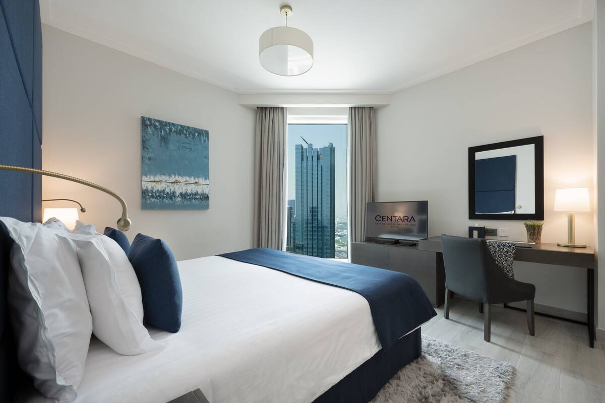 Una habitación de hotel moderna con una cama grande, un escritorio, una silla, un televisor y vistas a un paisaje urbano a través de la ventana.