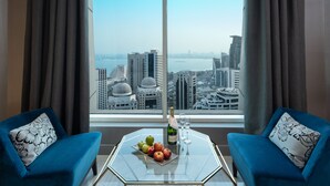 Una habitación con un ventanal grande, dos sillones azules, una mesa de vidrio con una botella de champán, una copa y un plato de fruta.