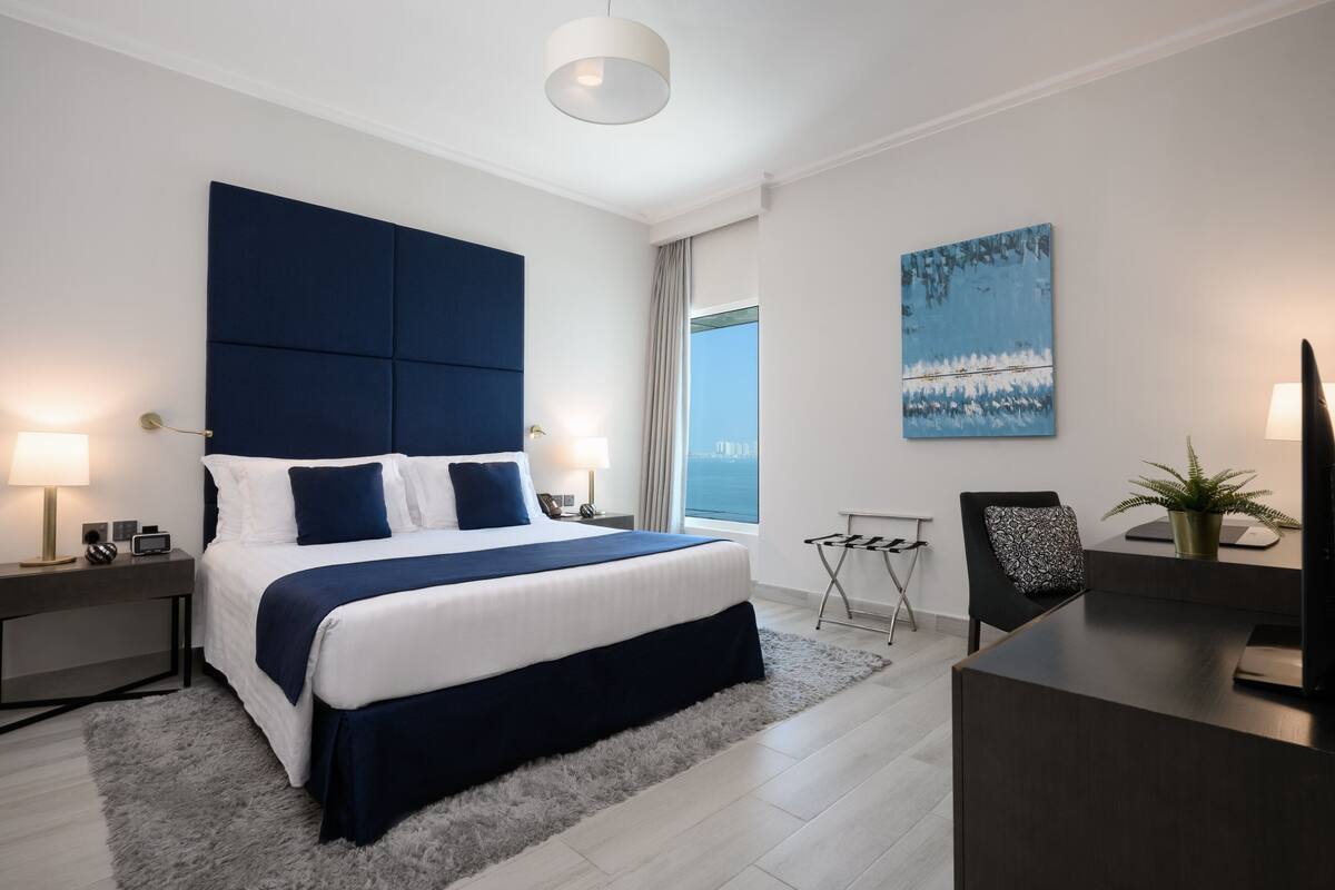 Habitación de hotel moderna con una cama grande, un escritorio, una silla, un televisor, una lámpara y una ventana con vistas.
