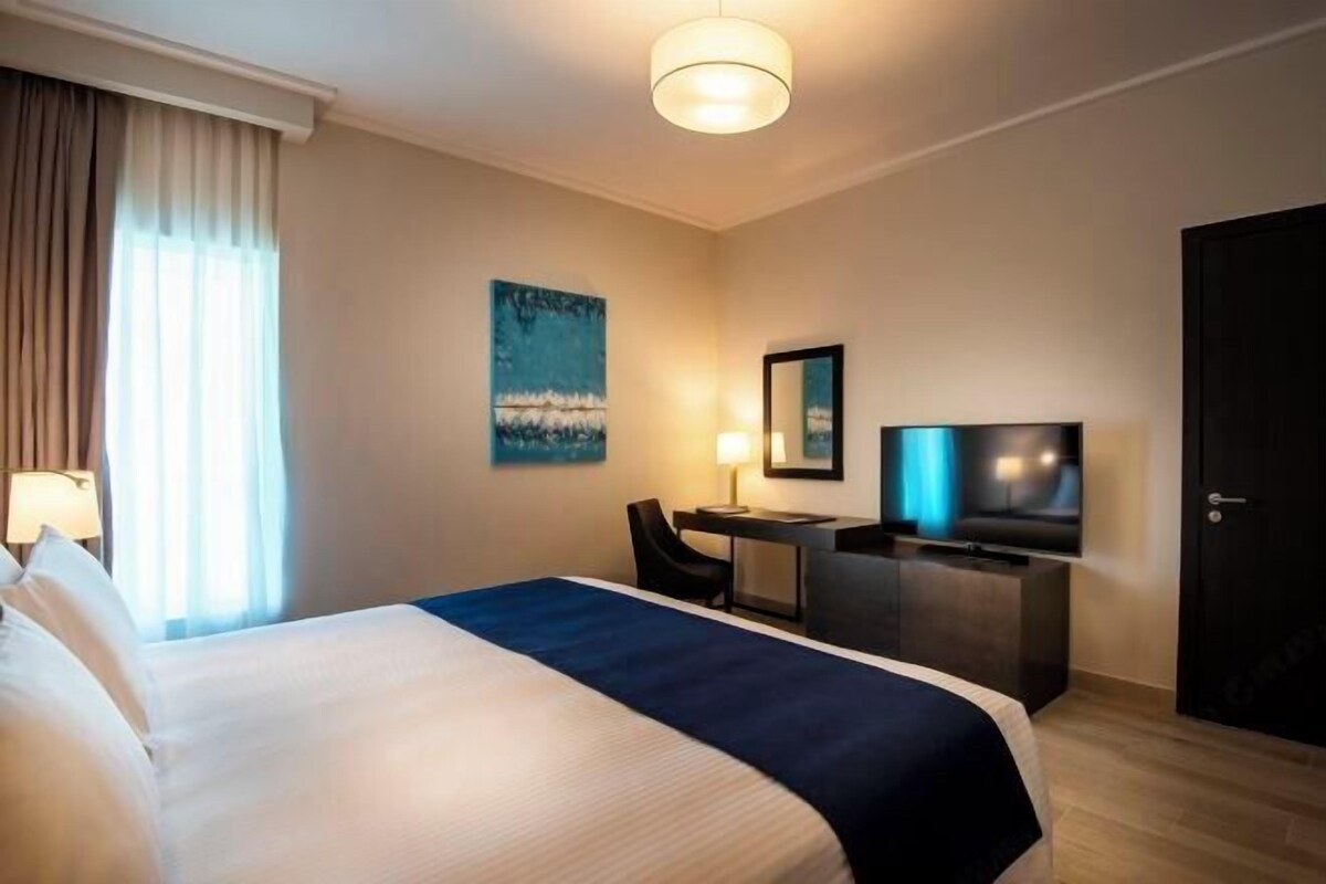 A hotel room with a large bed, a desk with a chair, a television, and a painting on the wall.
