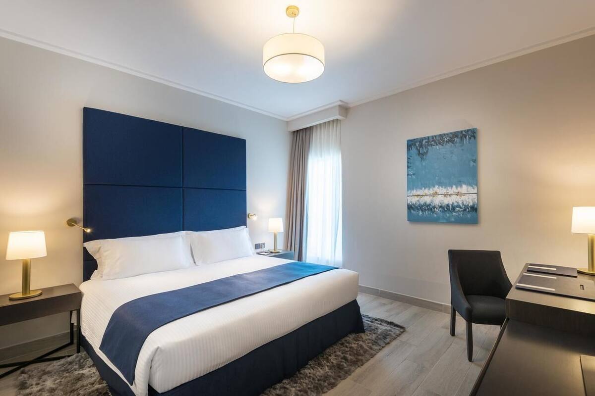 Una habitación de hotel moderna con una cama grande, un escritorio y una silla. Hay un cuadro en la pared y una ventana con cortinas.