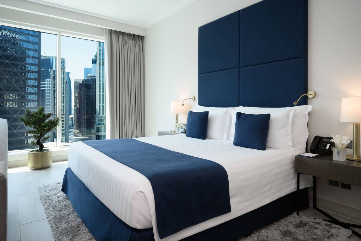 Una habitación de hotel moderna con una cama grande, vistas a la ciudad, una pequeña planta en maceta y una mesita de noche con lámpara y teléfono.