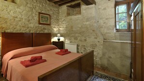 A rustic stone-walled bedroom with a wooden bed, a nightstand with a lamp, and a window with a view of greenery.