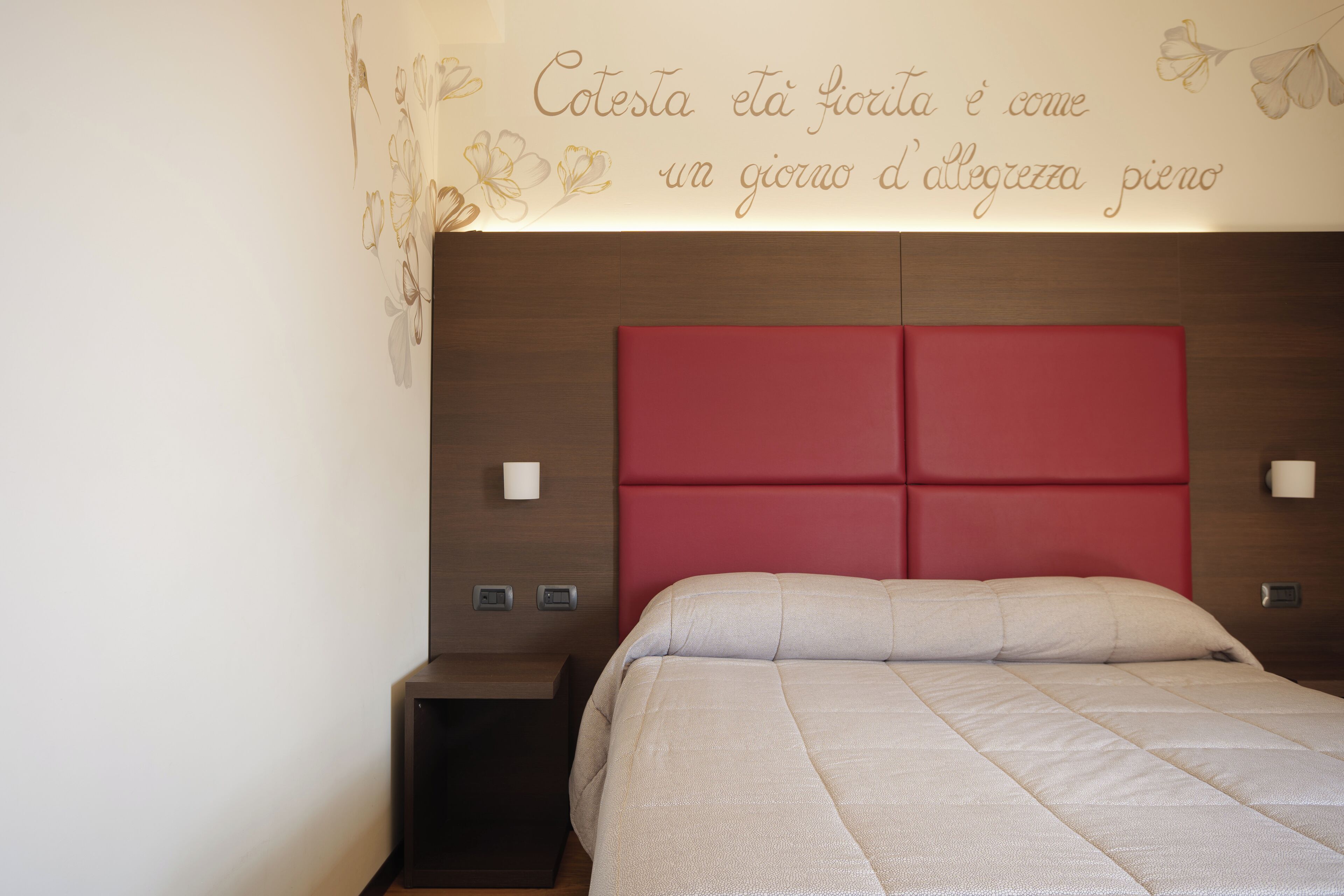 A bedroom with a red headboard, a beige bedspread, and a wall with a floral design and Italian text.