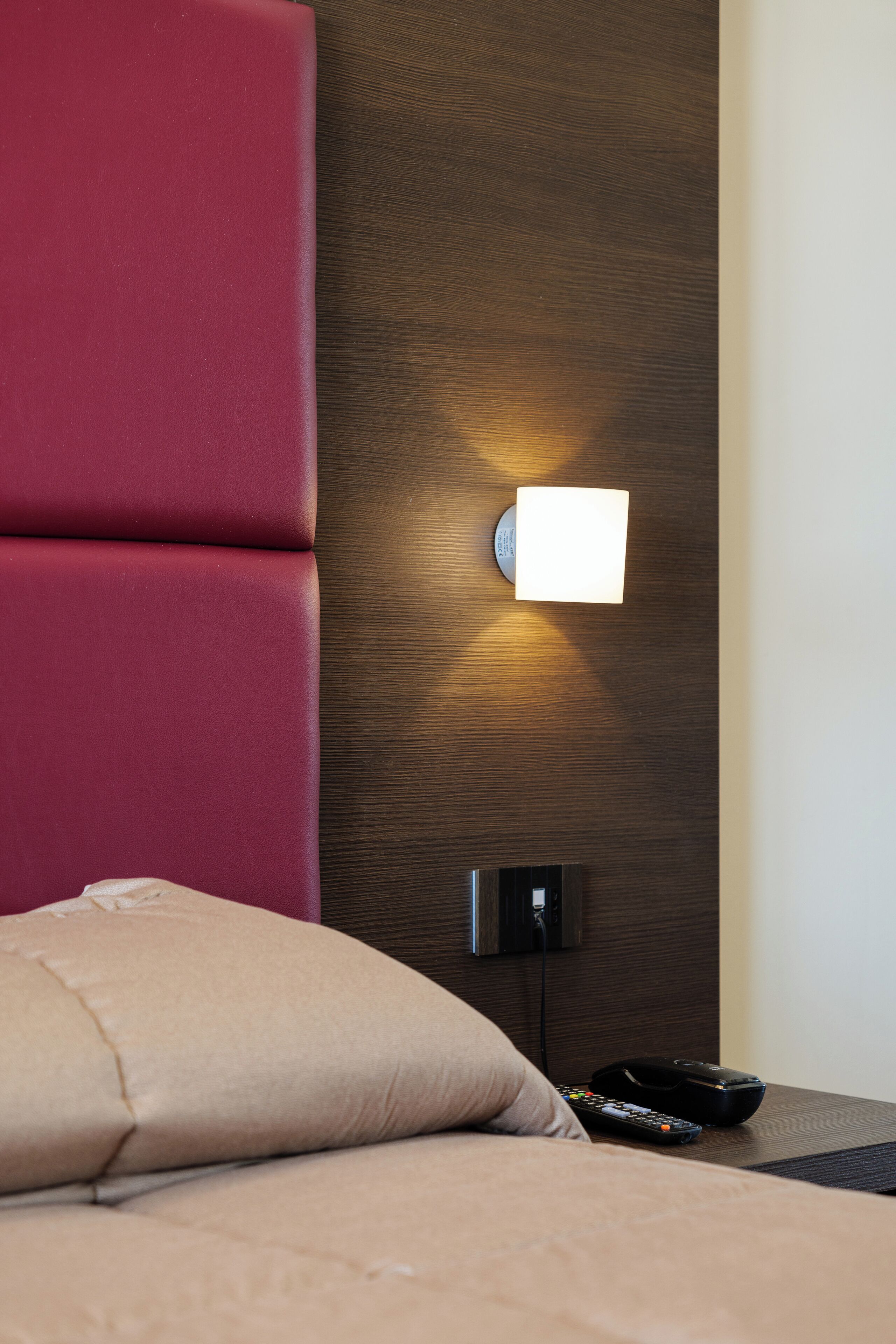 A modern hotel room with a red upholstered headboard, a bedside table with a remote control, and a wall-mounted light fixture.