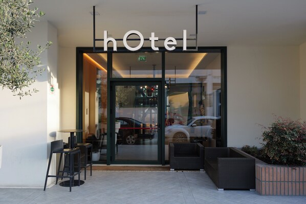 Entrada de un hotel con puerta de vidrio, mobiliario exterior y un cartel sobre la puerta.