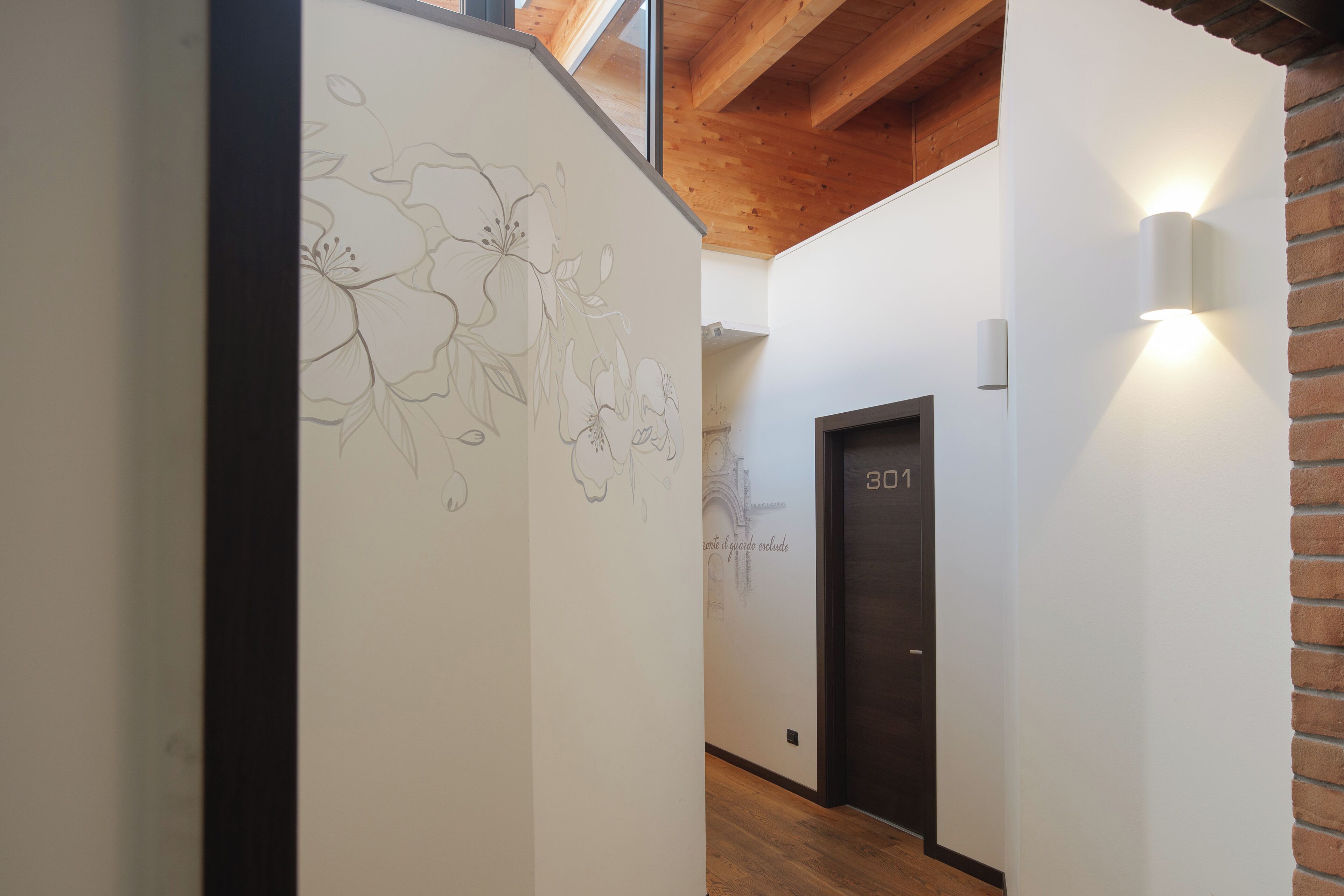 Un pasillo con techo de madera, una pared con dibujos florales y una puerta identificada con el número '301', además de una lámpara fijada a la pared.