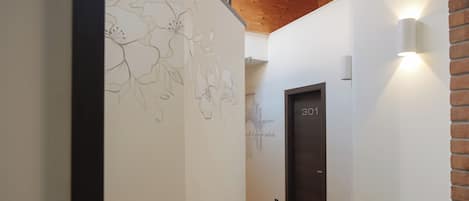 Un pasillo con techo de madera, una pared con dibujos florales y una puerta identificada con el número '301', además de una lámpara fijada a la pared.