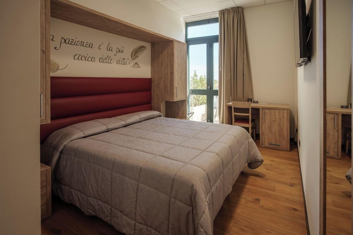 A bedroom with a bed, a desk, a chair, a TV, and a window with curtains.