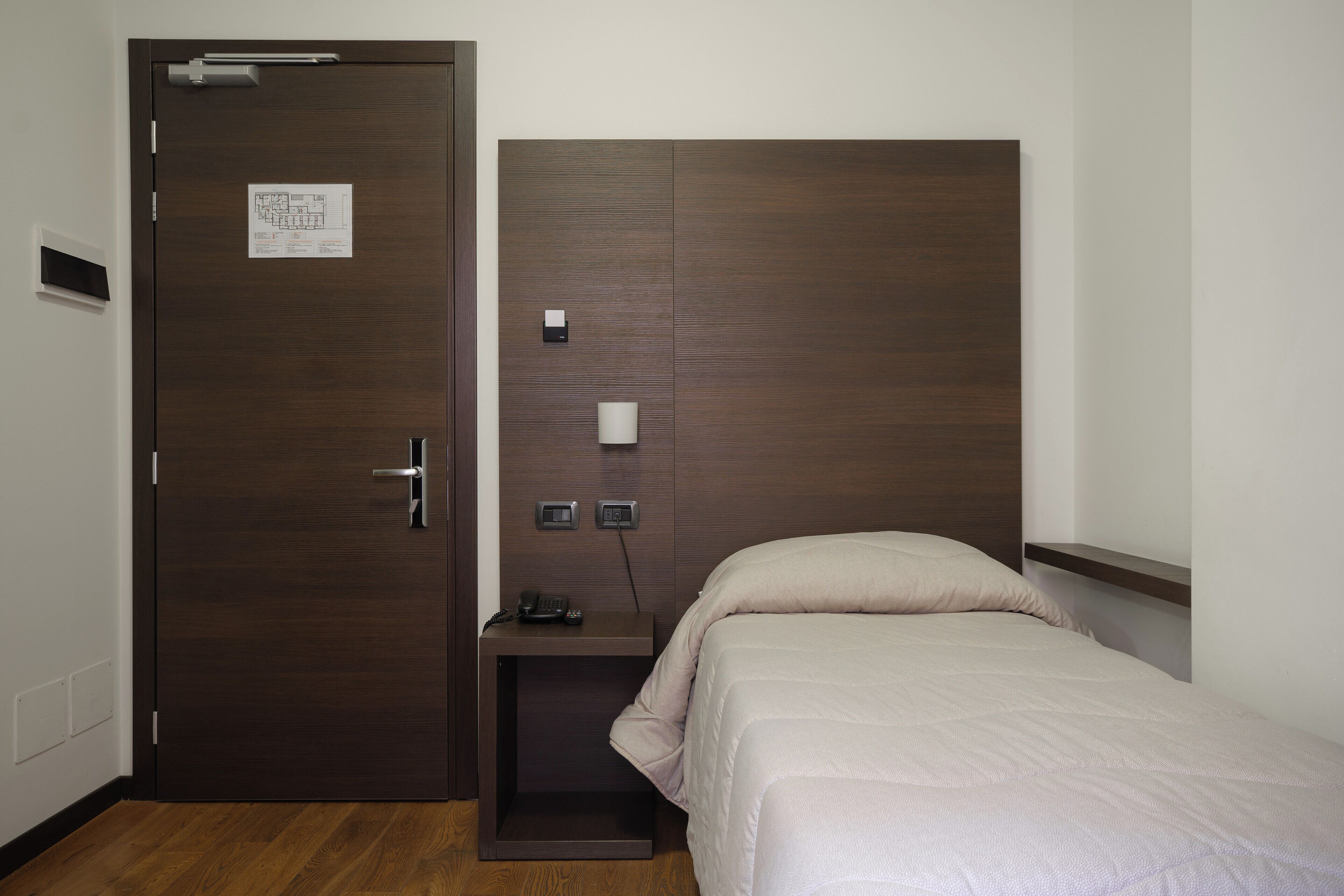 Une chambre d’hôtel avec une porte en bois, une table de chevet avec un téléphone et une lampe, et un lit recouvert d’une couette beige.