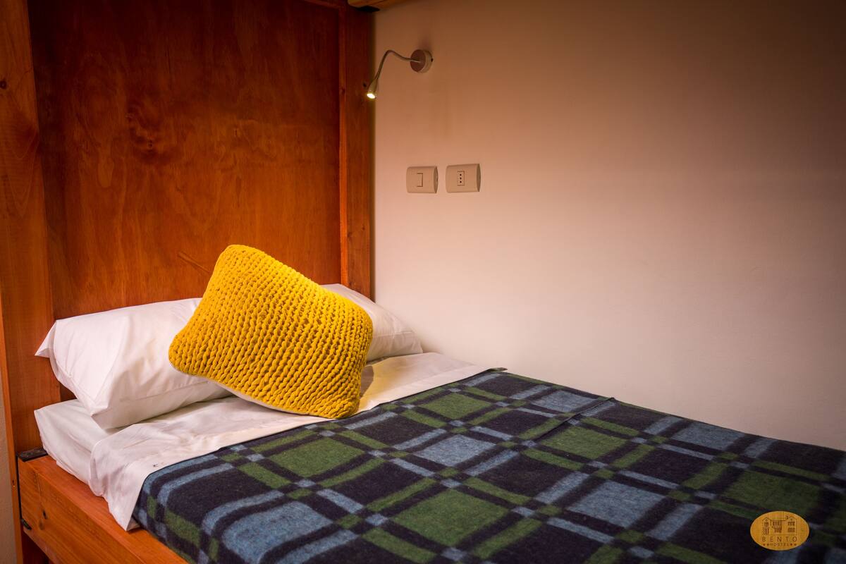A single bed with a yellow knitted pillow, a plaid blanket, and a wooden headboard.