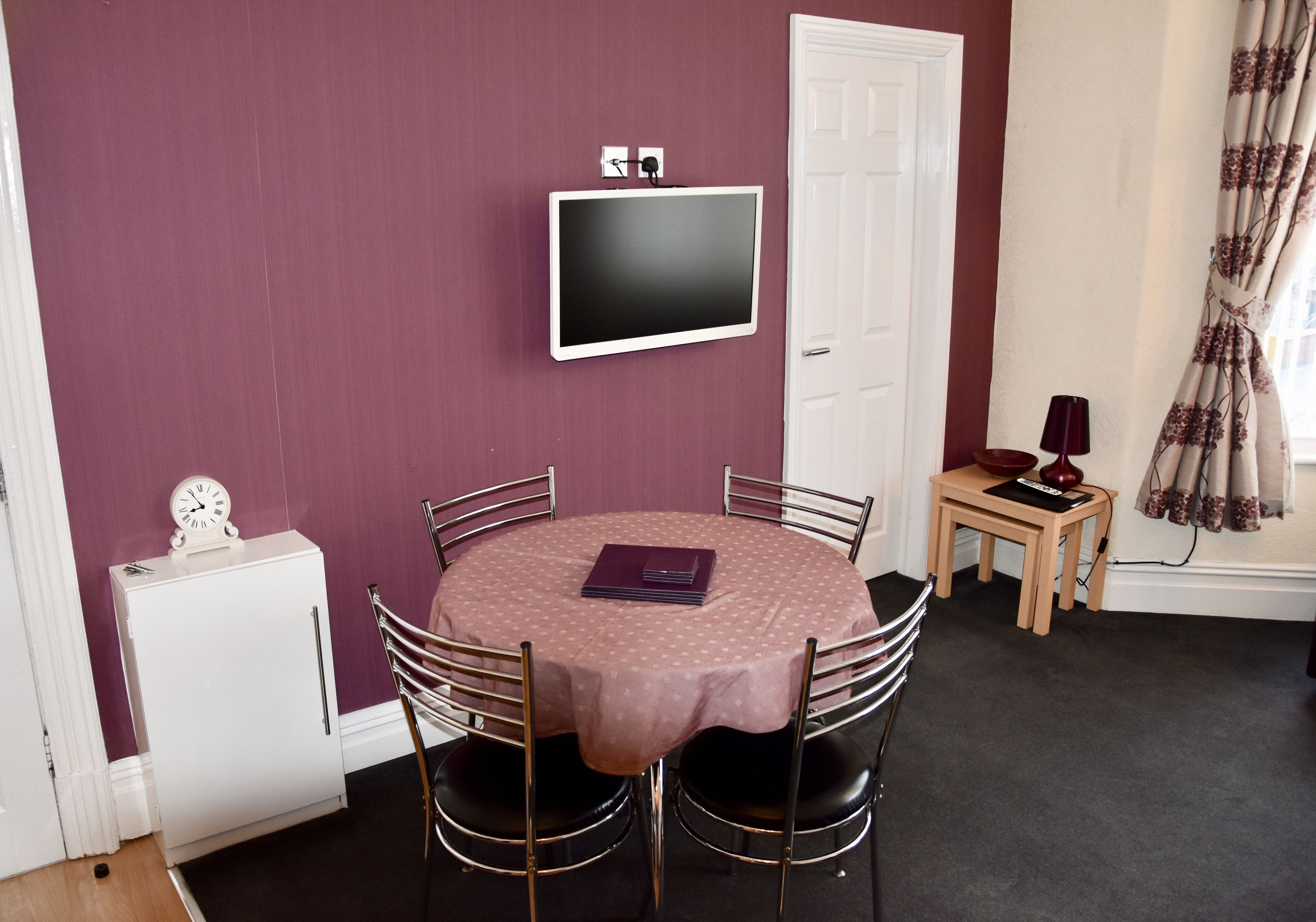 A dining area with a round table, chairs, a clock, a television, and a small cabinet.
