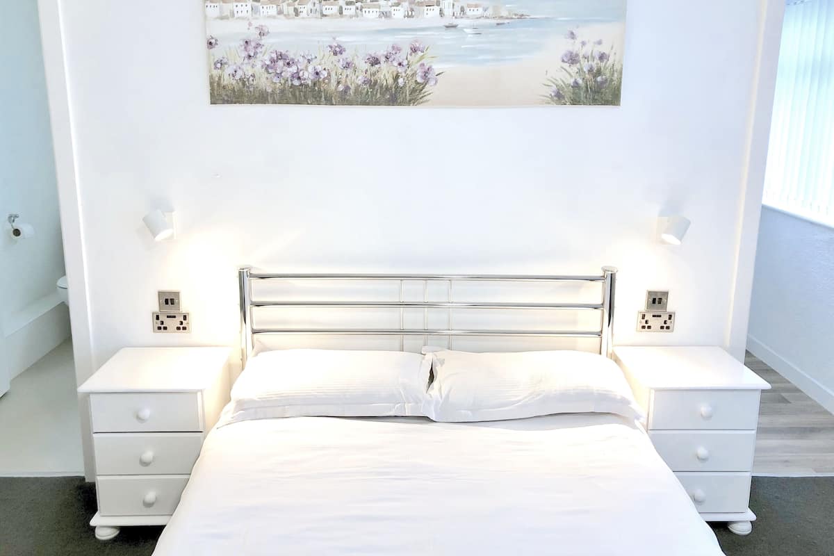 A bedroom with a bed, two bedside tables, a painting of a coastal scene, and a window with blinds.