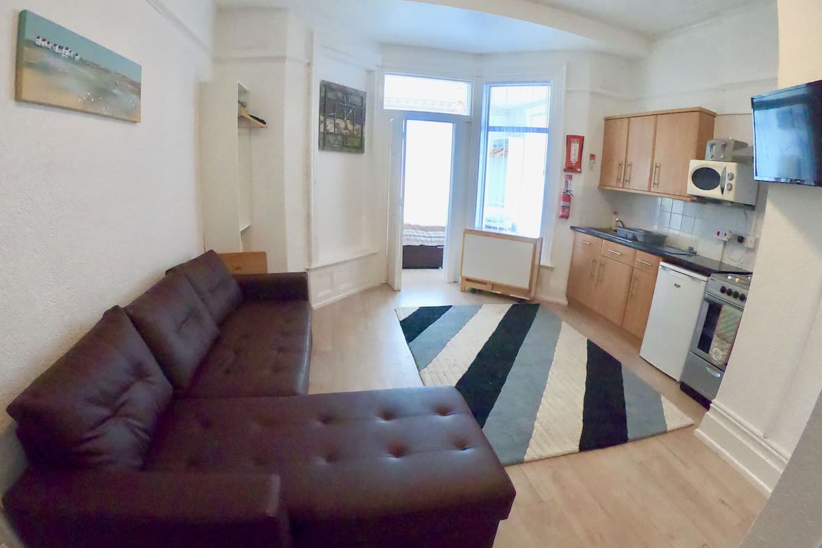 A living room with a dark brown sofa, a kitchen with wooden cabinets, a microwave, and a stove.
