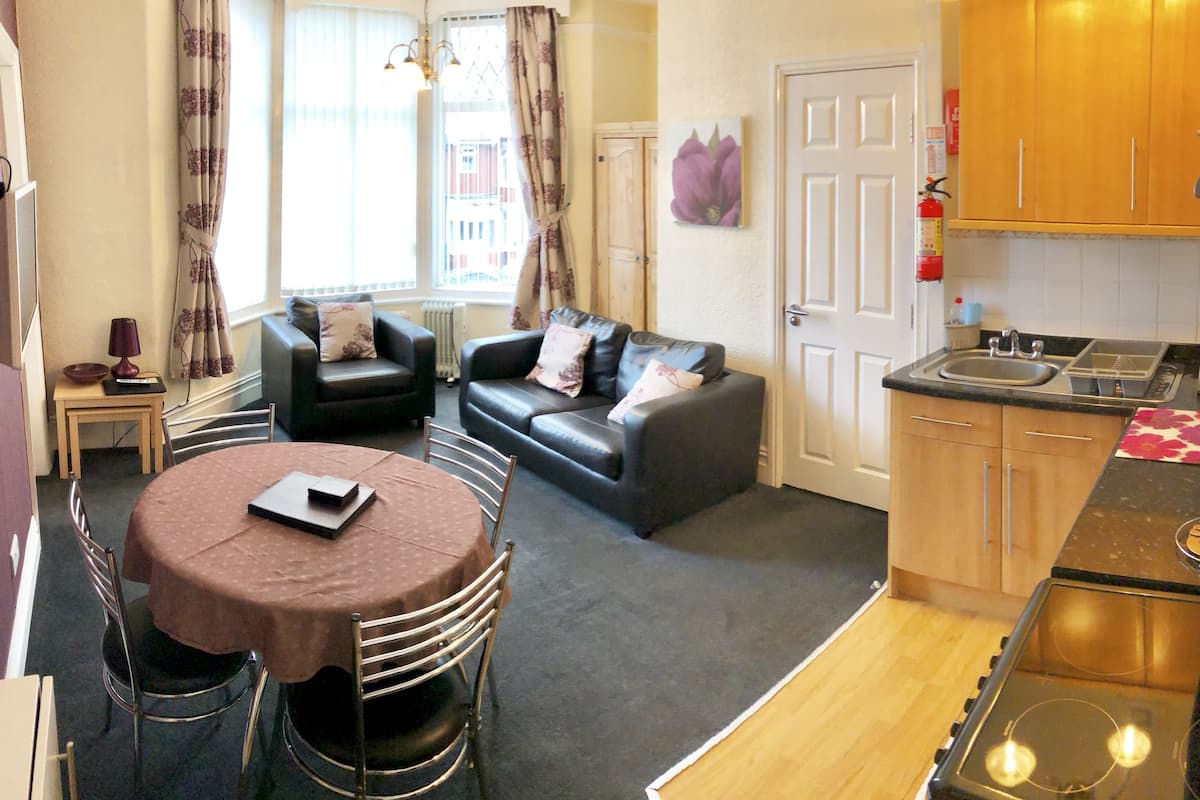 A compact living space with a dining area, a sofa, a kitchen with a stove and sink, and a window with curtains.
