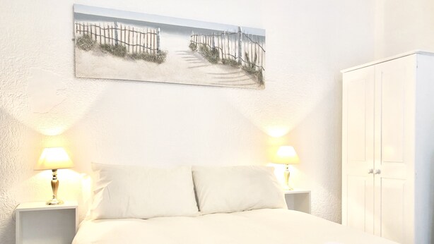 Una cama bien hecha con sábanas blancas, dos lámparas de noche, un cuadro colgado en la pared que muestra una playa y un armario blanco.