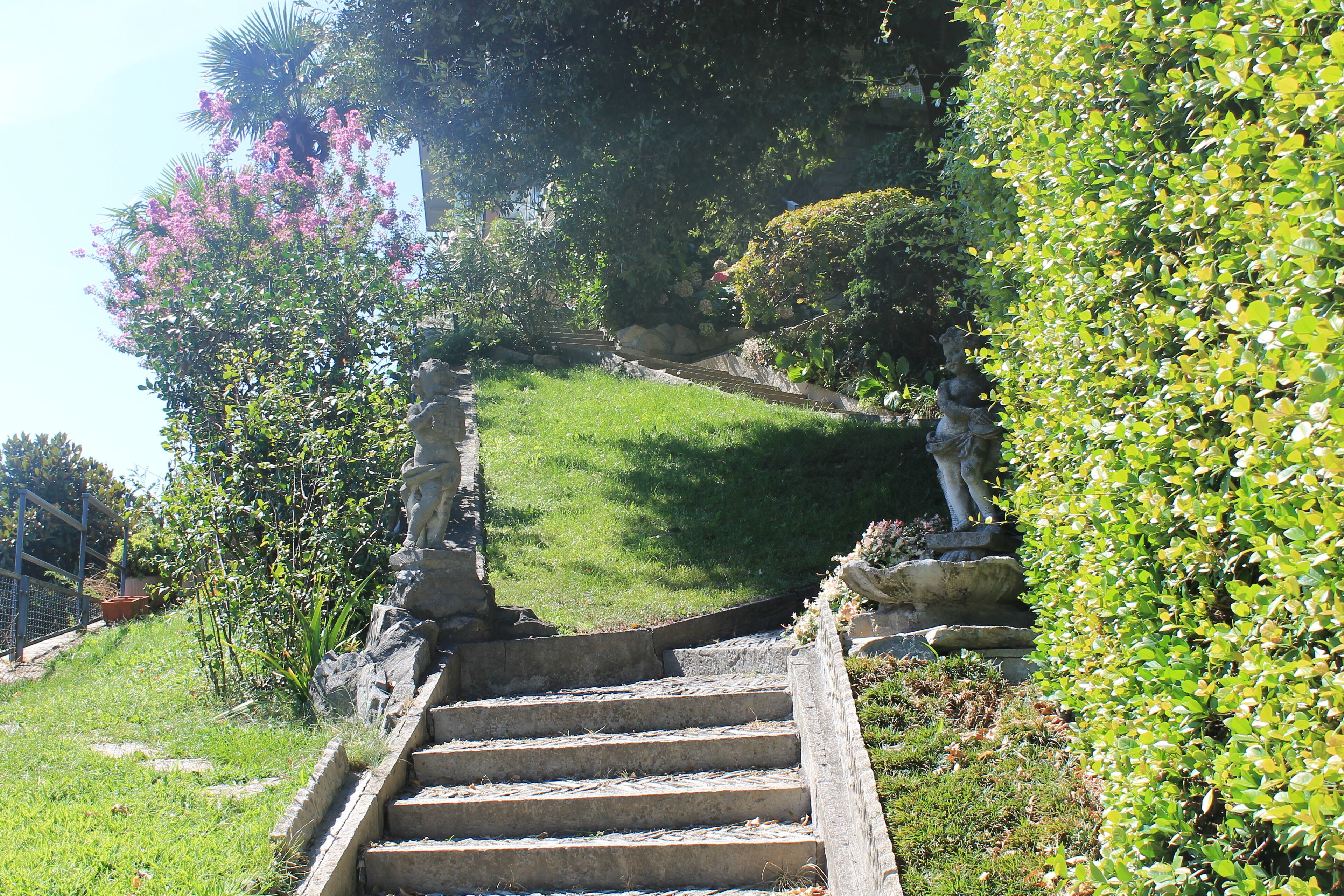 Eine steile Steintreppe führt über einen grasbewachsenen Hang, umgeben von üppigem Grün und einer Statue.