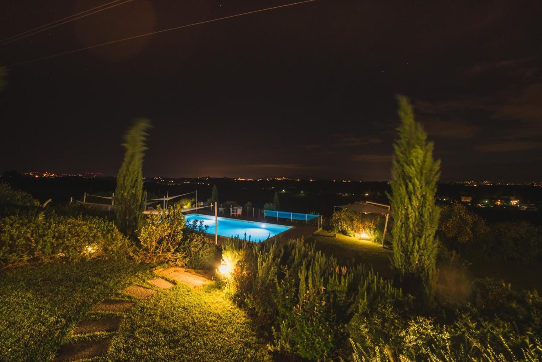 Une vue nocturne d’une piscine, entourée de végétation, avec les lumières de la ville qui scintillent au loin.