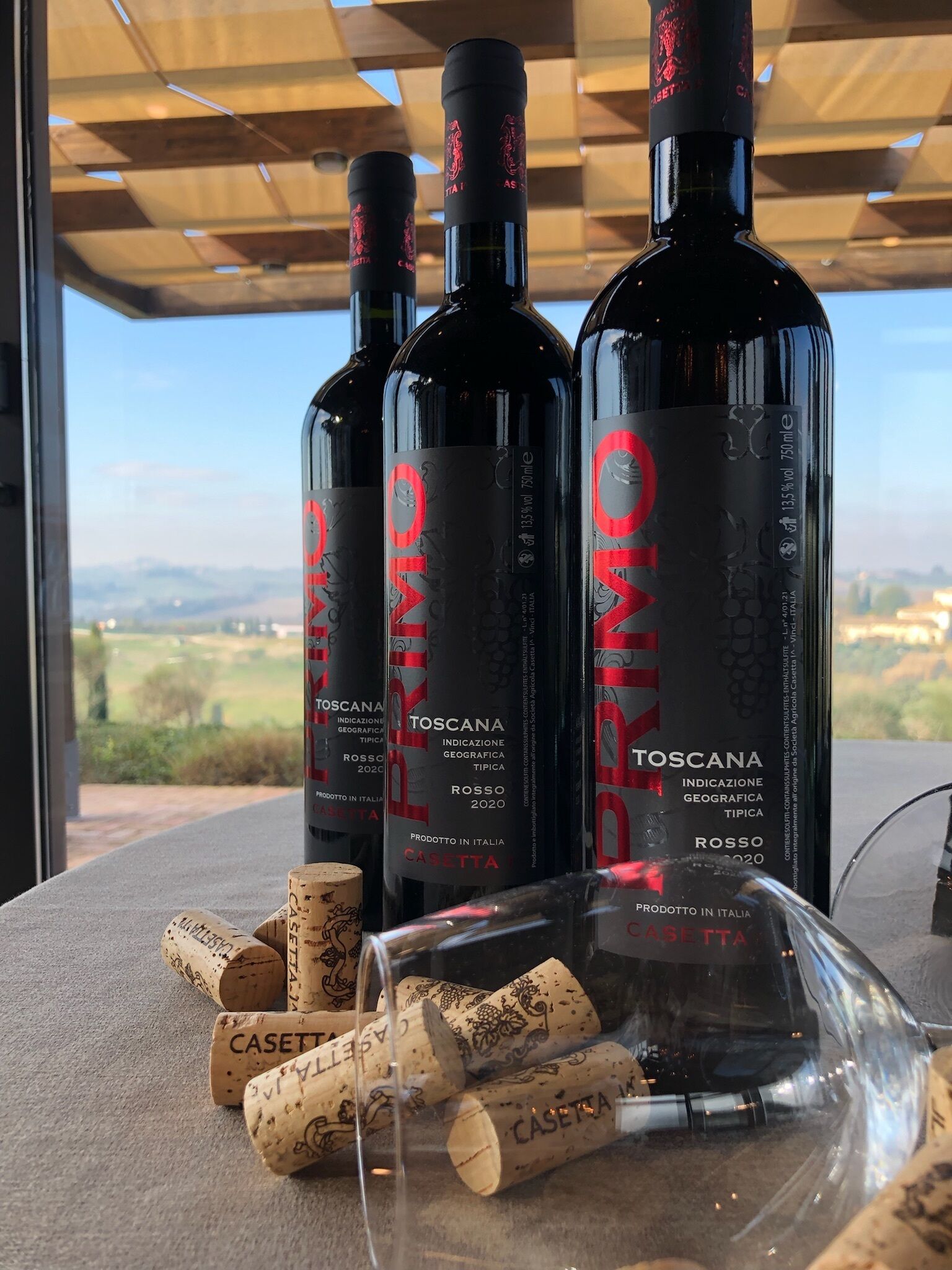 Tre bottiglie di vino Toscana Rosso 2020 e diversi tappi su un tavolo con vista sulla campagna.