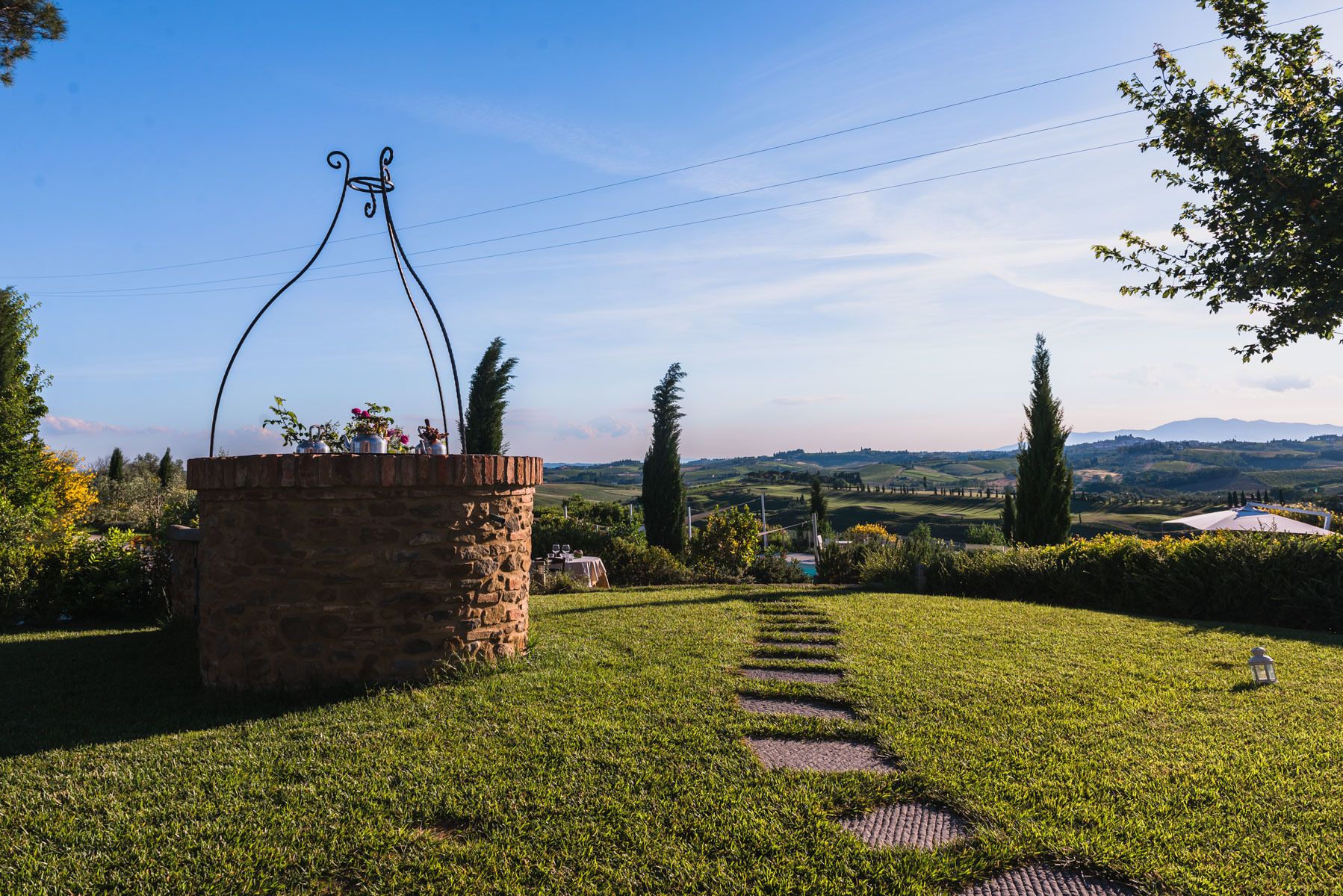 A stone structure with a metal archway, surrounded by a grassy area with a clear path leading to a distant view of rolling hills and a body of water.