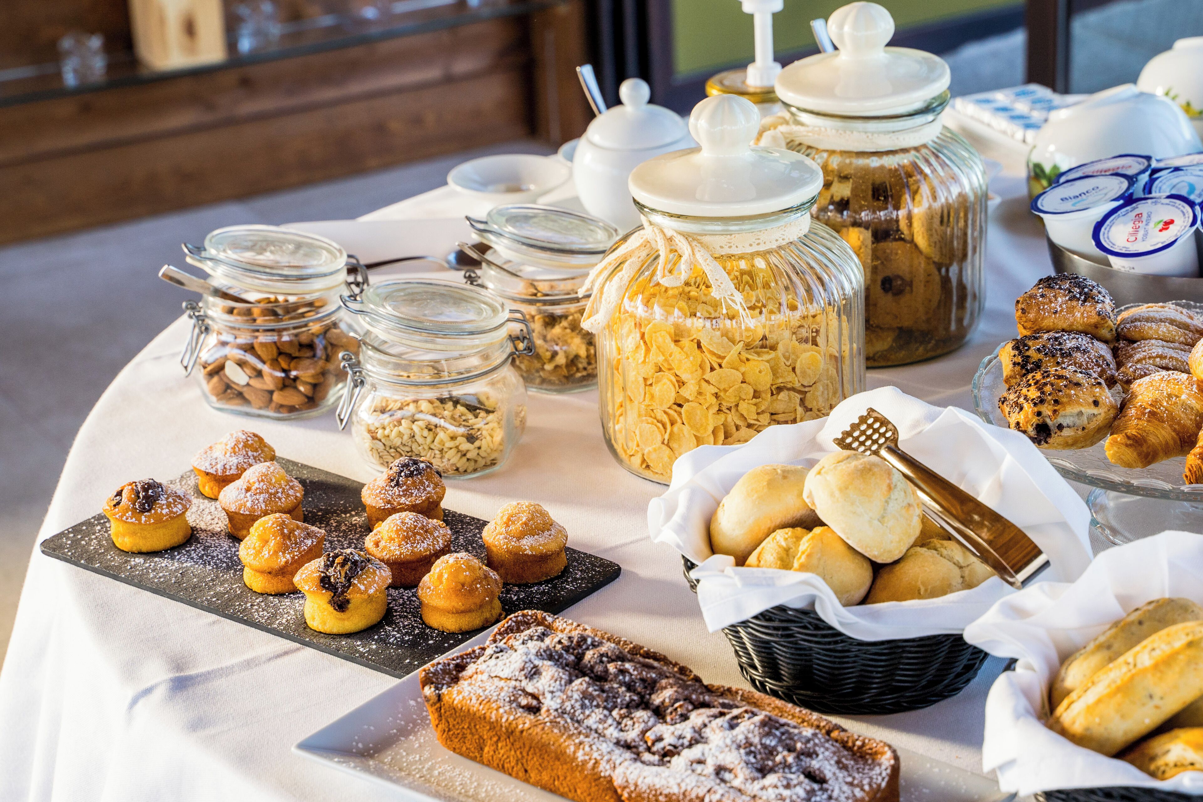 Uma mesa de buffet com diversos produtos de panificação, incluindo doces, muffins e pão, dispostos sobre uma toalha de mesa branca.