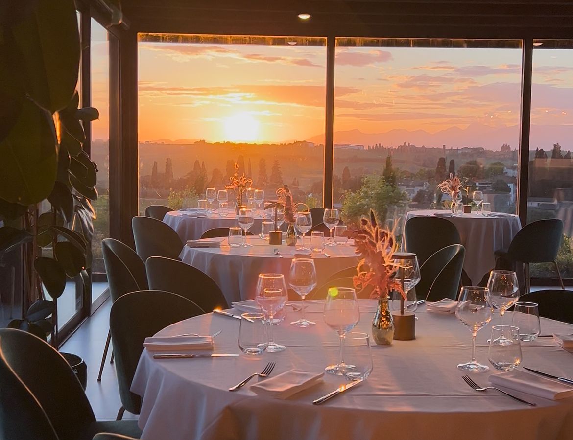 A restaurant with tables set for dining, large windows with a sunset view, and indoor plants.