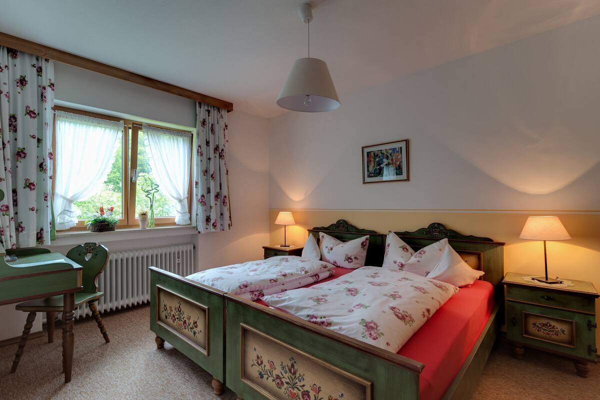 Ein Schlafzimmer mit einem Bett, zwei Nachttischlampen, einem Fenster mit Vorhängen, einer Heizung, einem Schreibtisch mit Stuhl und einem gerahmten Bild an der Wand.