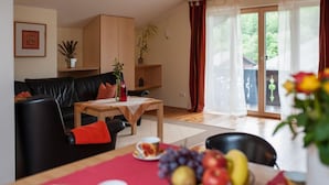Una sala de estar con un sofá de cuero negro, una mesa de centro de madera y un comedor con mantel rojo. Hay plantas en estanterías y un ventanal corredero que da a un área exterior.