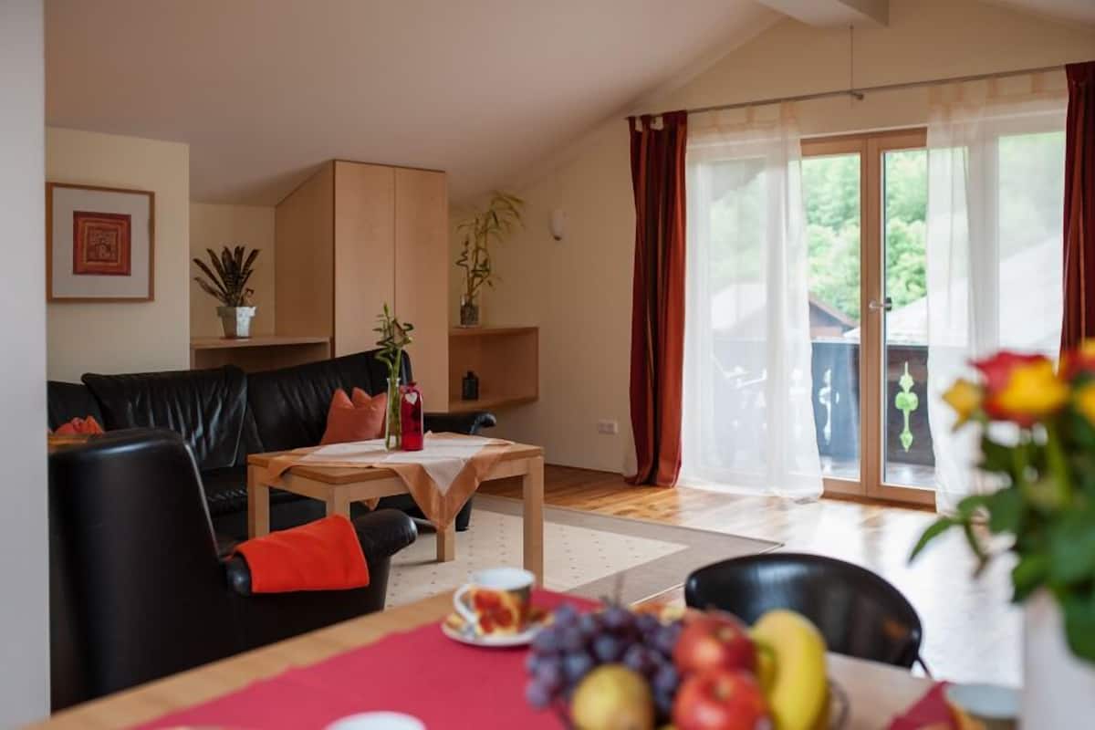 Ein Wohnzimmer mit einer schwarzen Ledersofa, einem Holztisch und einem Essbereich mit roter Tischdecke. Pflanzen stehen auf Regalen, und eine Glastür öffnet sich zu einem Außenbereich.
