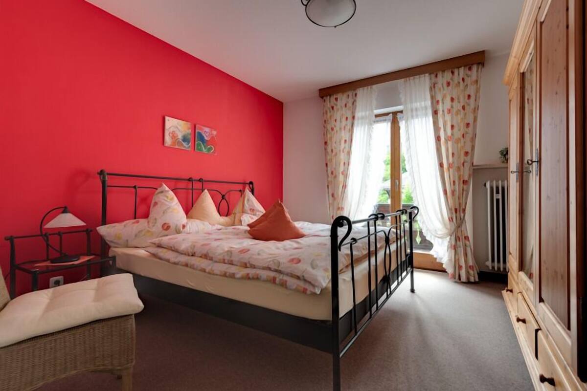 Ein Schlafzimmer mit Bett, Nachttisch, Lampe, Sessel und einem Fenster mit Vorhängen.