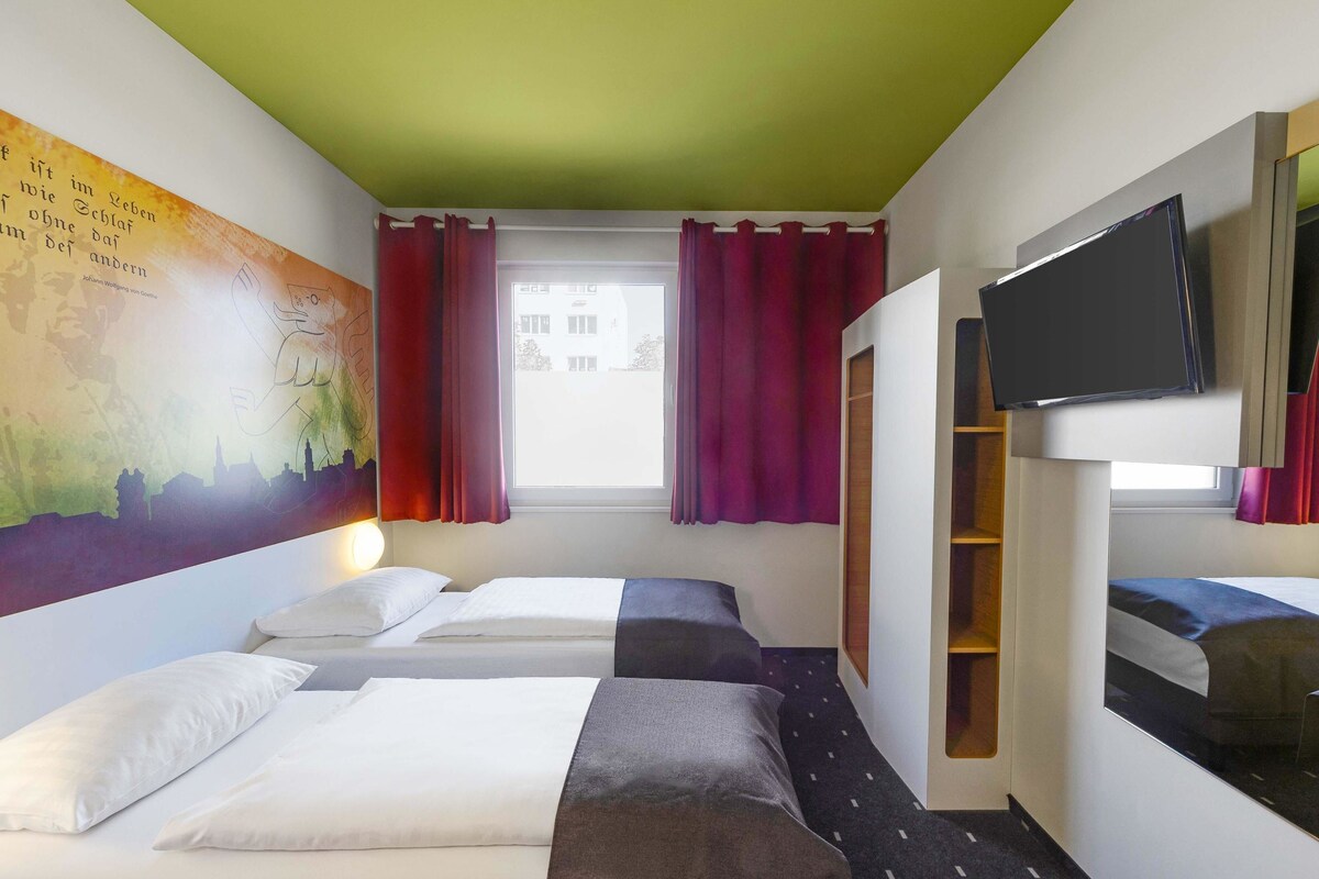 A hotel room with two beds, a TV, a mural, and a window with curtains.