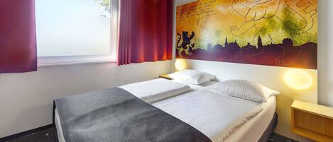 A hotel room with a bed, a window with curtains, a wall with a cityscape painting, and a nightstand with a lamp.