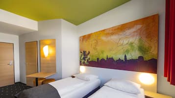 Quarto de hotel com duas camas de solteiro, uma escrivaninha de madeira, uma luminária fixada na parede e um quadro abstrato grande.