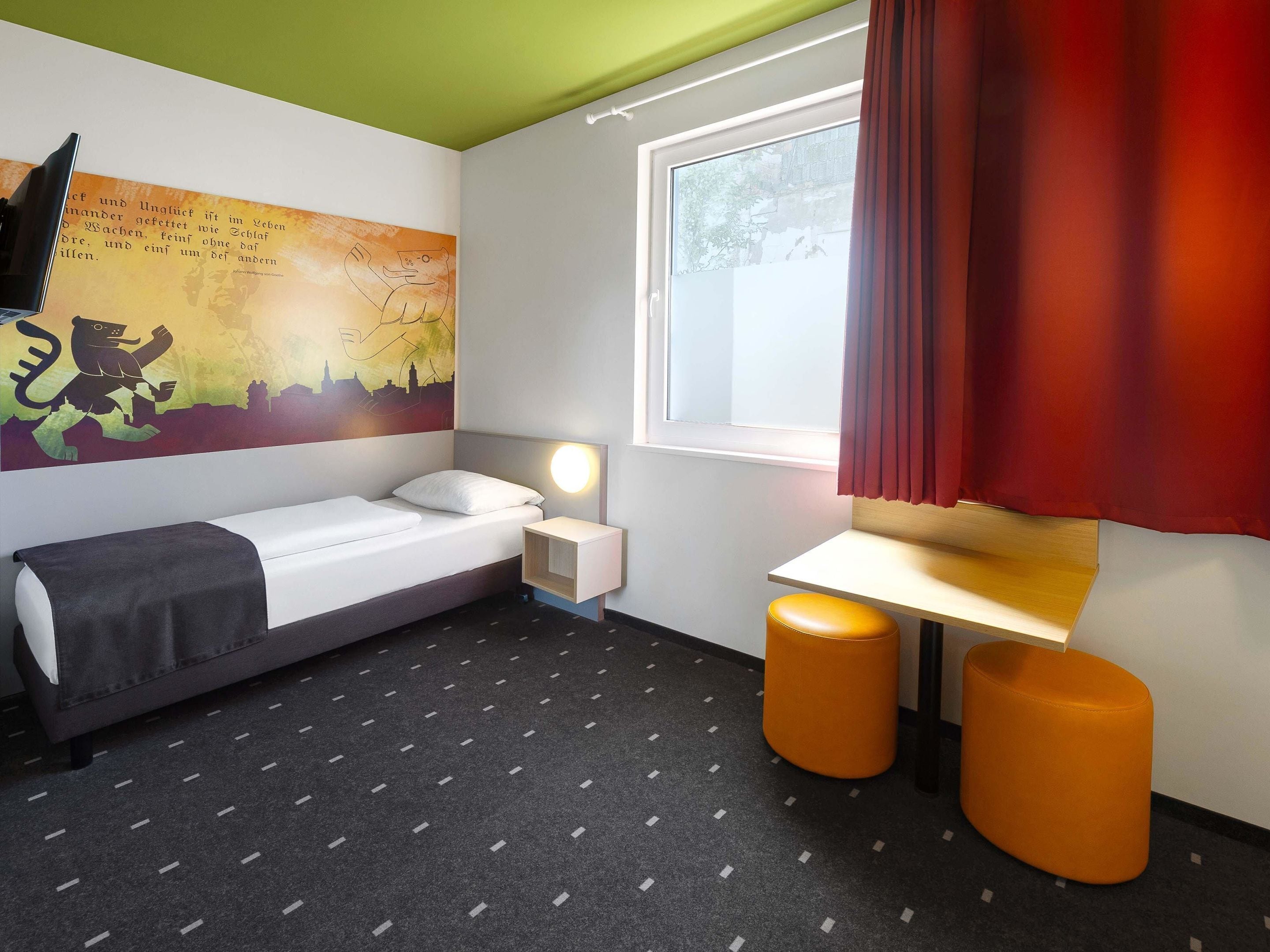 Quarto de hotel com uma cama de solteiro, uma mesa de cabeceira, uma escrivaninha, dois banquinhos laranja, uma janela com cortinas vermelhas e um mural com vista para a cidade.