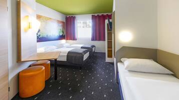 Habitación de hotel con una cama, dos camas individuales, un escritorio, un reposapiés naranja, un cuadro en la pared y una ventana con cortinas rojas.