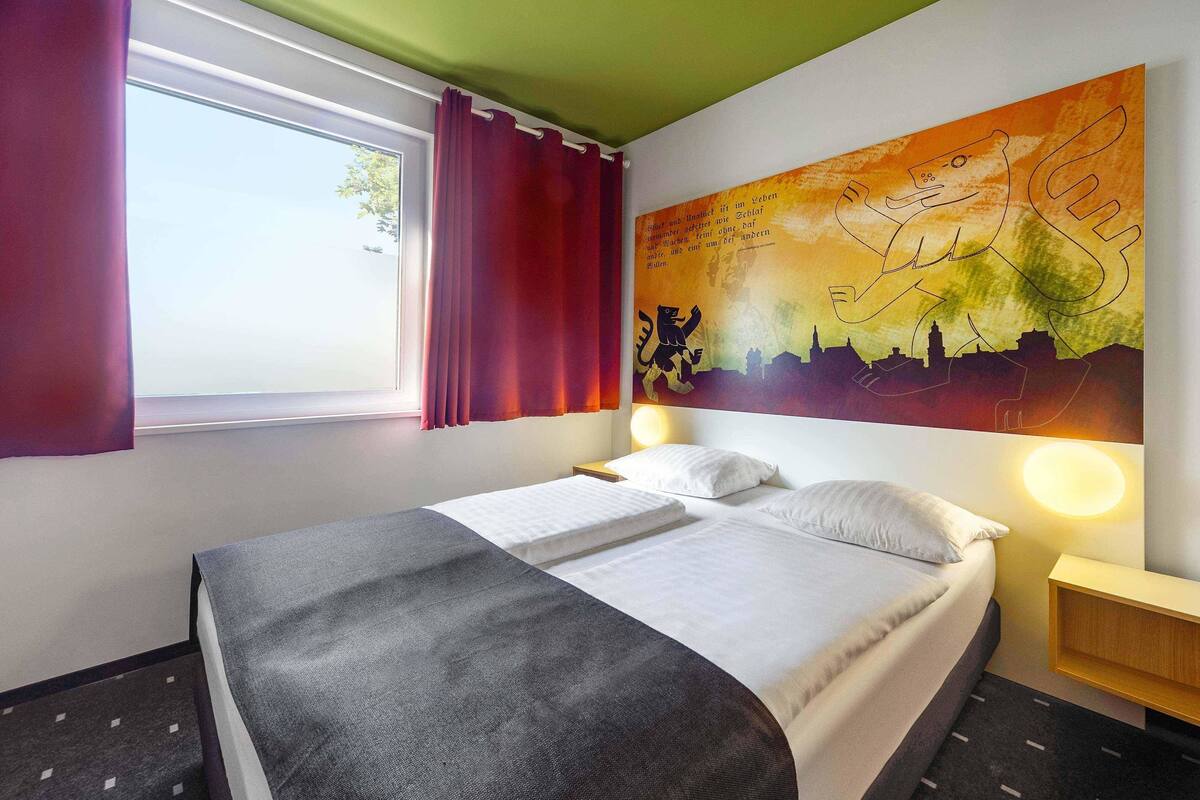 A hotel room with a bed, a window with curtains, a wall with a cityscape painting, and a nightstand with a lamp.