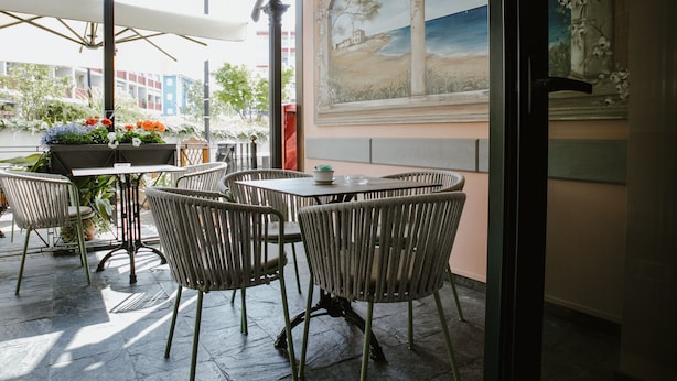 Une terrasse extérieure avec des chaises et une table en osier, une fresque représentant un paysage côtier, et une vue sur des bâtiments et des arbres.