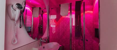 Un baño moderno con ducha iluminada en rosa, un secador de pelo y un lavabo.