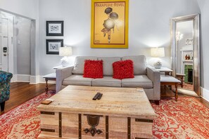 A living room with a grey sofa, red throw pillows, a wooden coffee table, and a red patterned rug. There are framed pictures on the wall and a large poster above the sofa. - Downtown Digs for Four, Walk Everywhere by Lucky Savannah (Savannah)