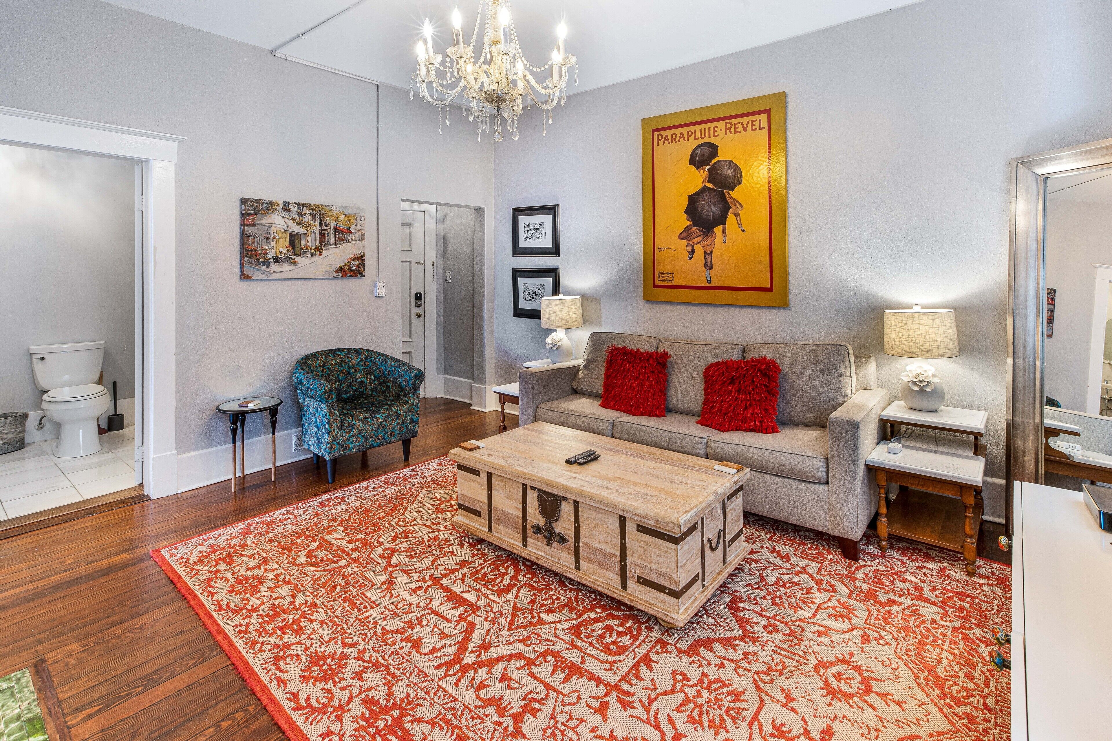 A living room with a wooden coffee table, a grey sofa with red cushions, a blue armchair, a chandelier, and a red patterned rug.